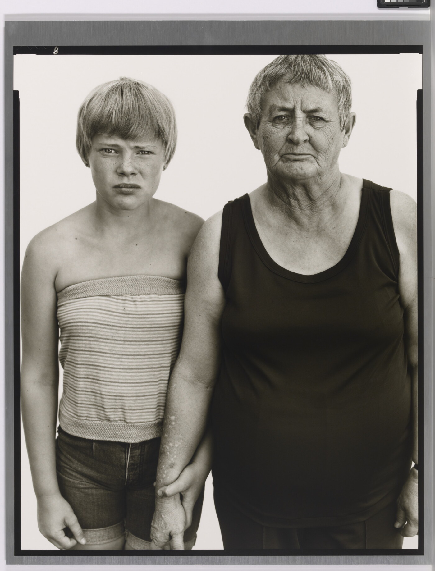 A black-and-white portrait photograph of a young White girl with short blonde hair wearing a tube top, and an older White woman with short gray hair wearing a black tank top.