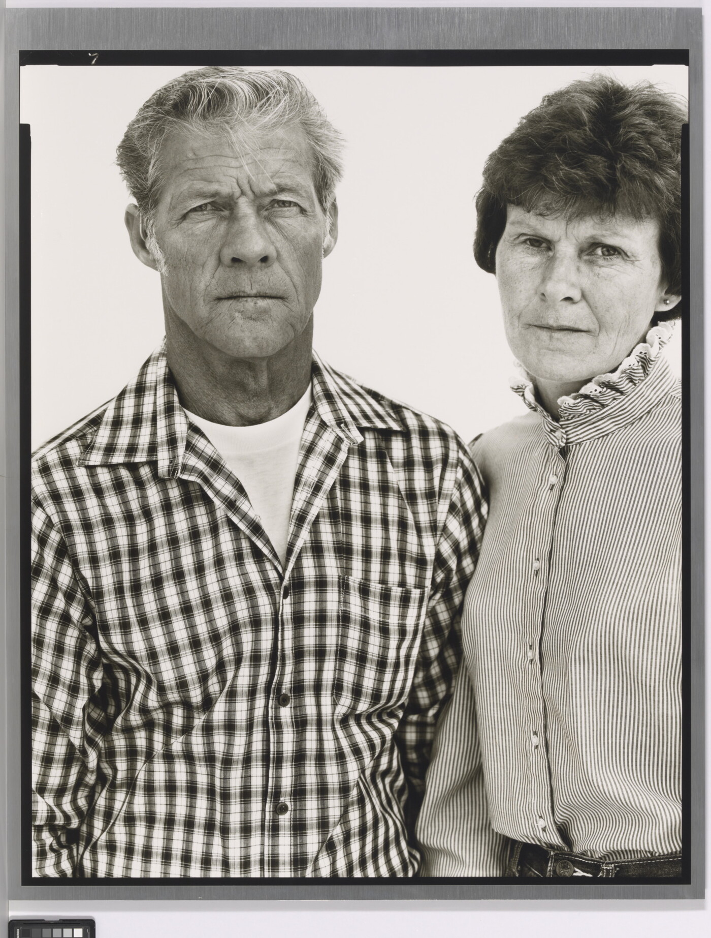 A black-and-white portrait photograph of a middle-aged White man wearing a checked button-down shirt and a middle-aged White woman in a striped high-collar blouse standing shoulder to shoulder.