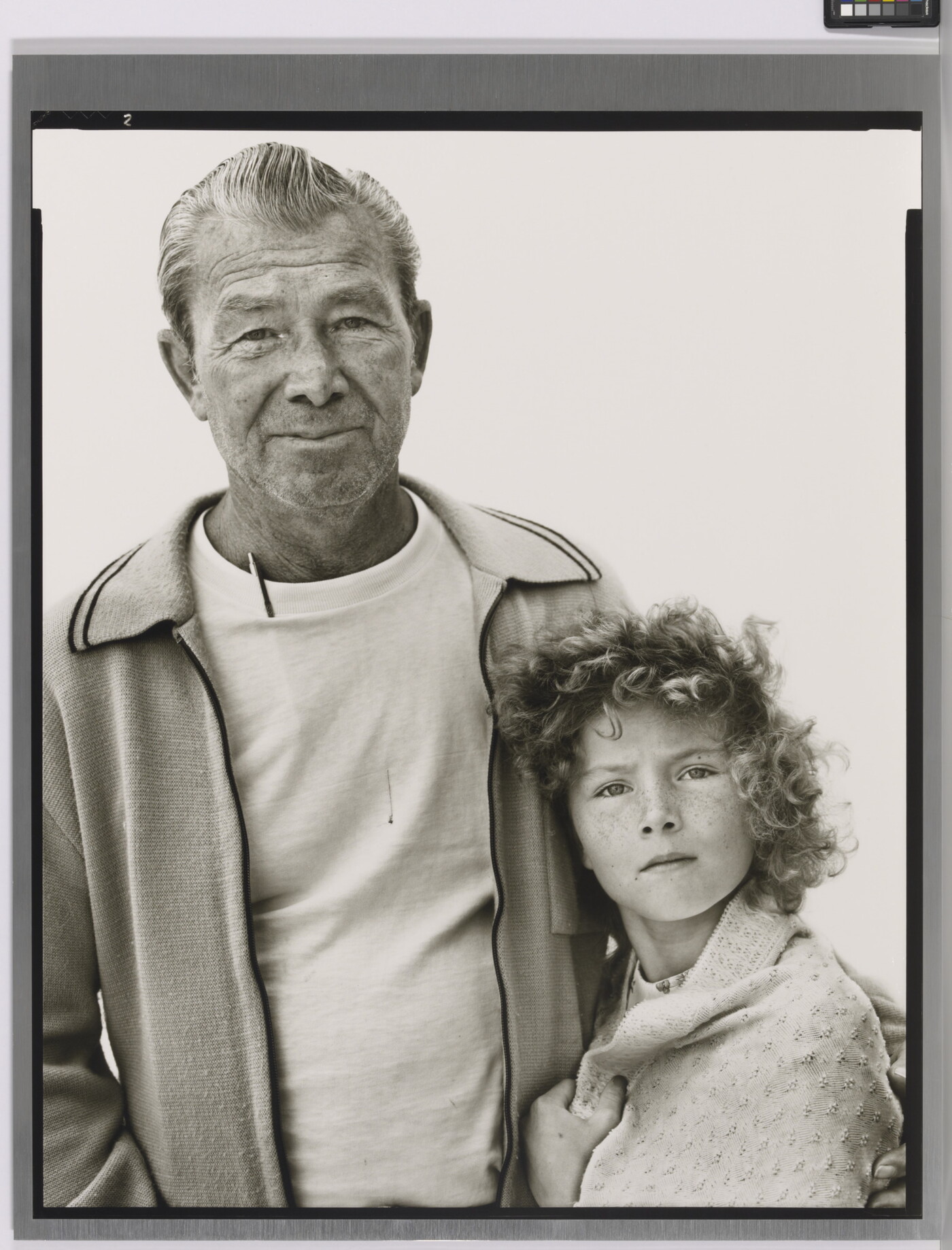 A black-and-white portrait photograph of an older White man in a t-shirt and cardigan with his arm around a young White girl with curly hair and a blanket wrapped around her shoulders.