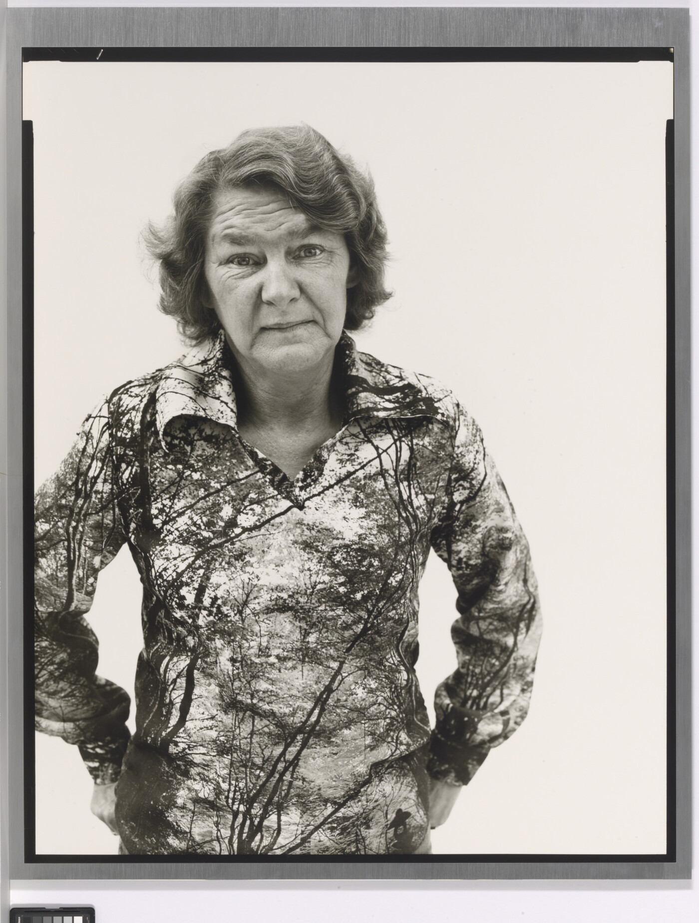 A black-and-white portrait photograph of an older White woman with wavy hair wearing a patterned polo shirt, hands hooked into her back pants pockets.