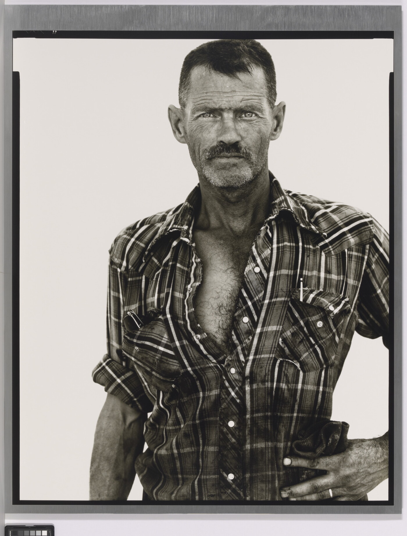 A black-and-white portrait photograph of a middle-aged White man with scruffy facial hair wearing a plaid button-down shirt standing with one hand on his hip.