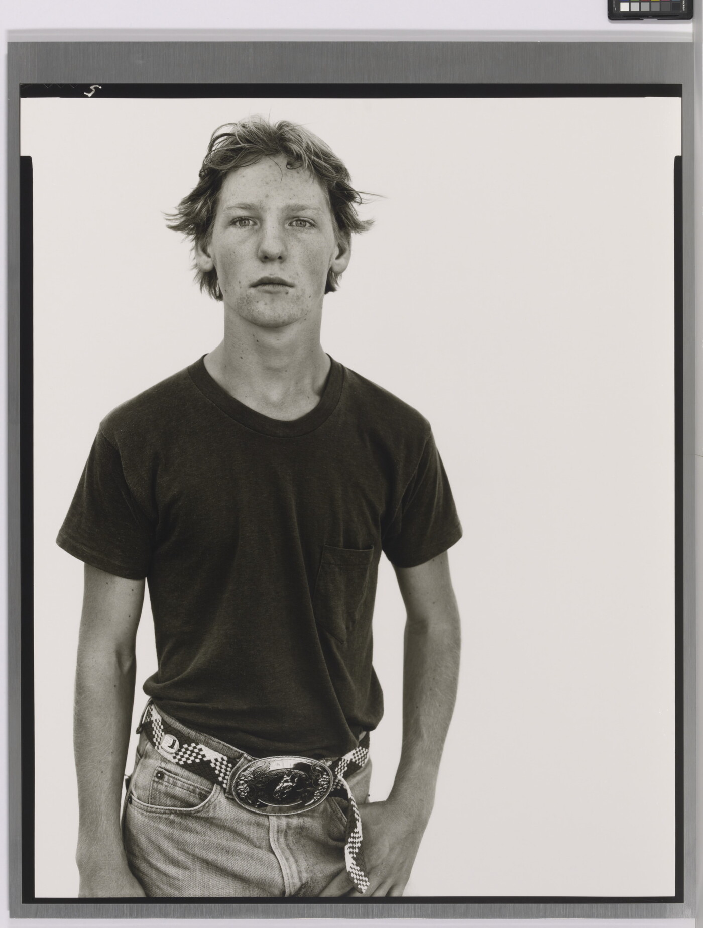 A black-and-white portrait photograph of a White teenage boy in a dark t-shirt and belted jeans with a large belt buckle, one hand hooked into his jeans pocket.