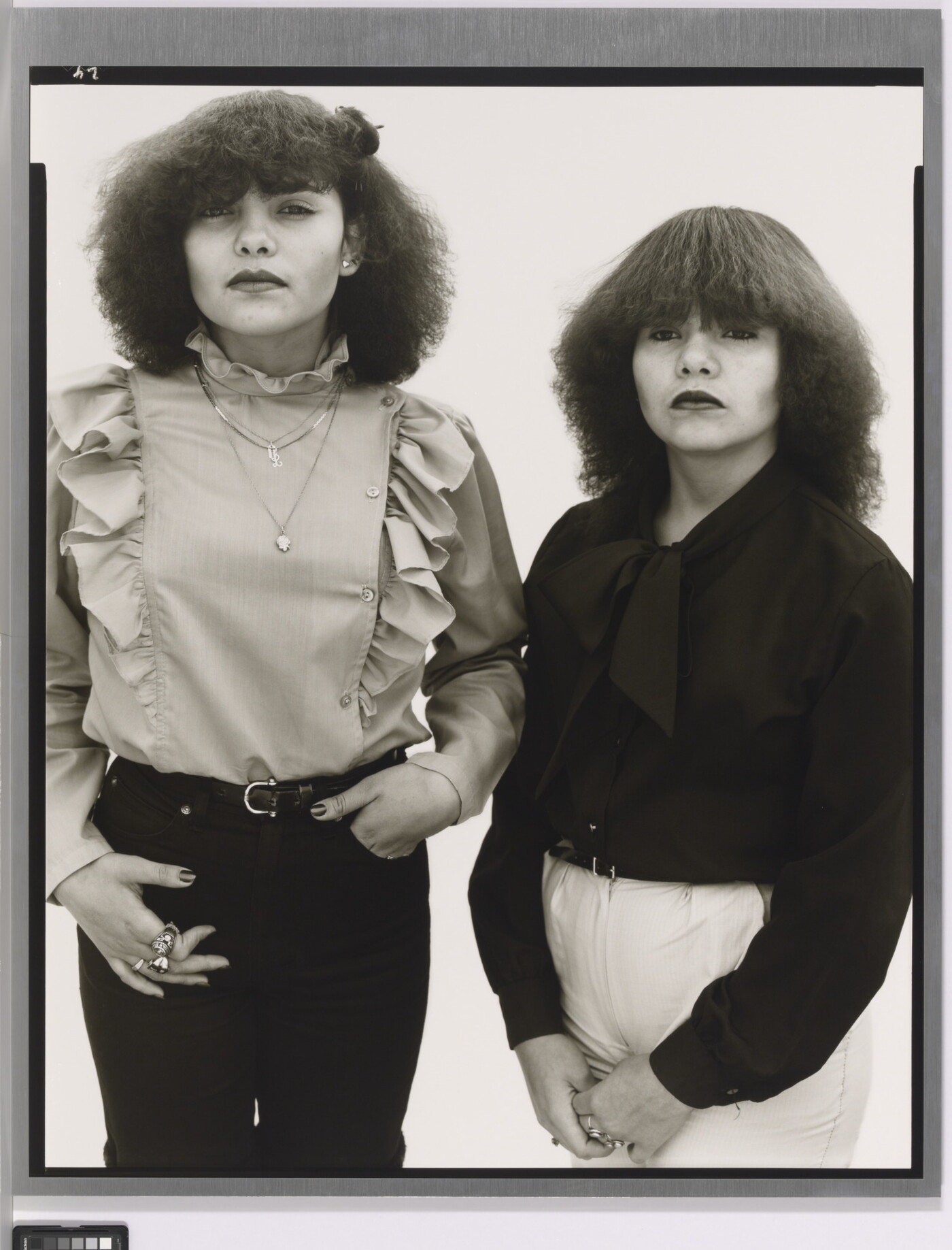 A black-and-white portrait photograph of two medium-skinned young women, both with teased dark hair and wearing ruffled blouses.