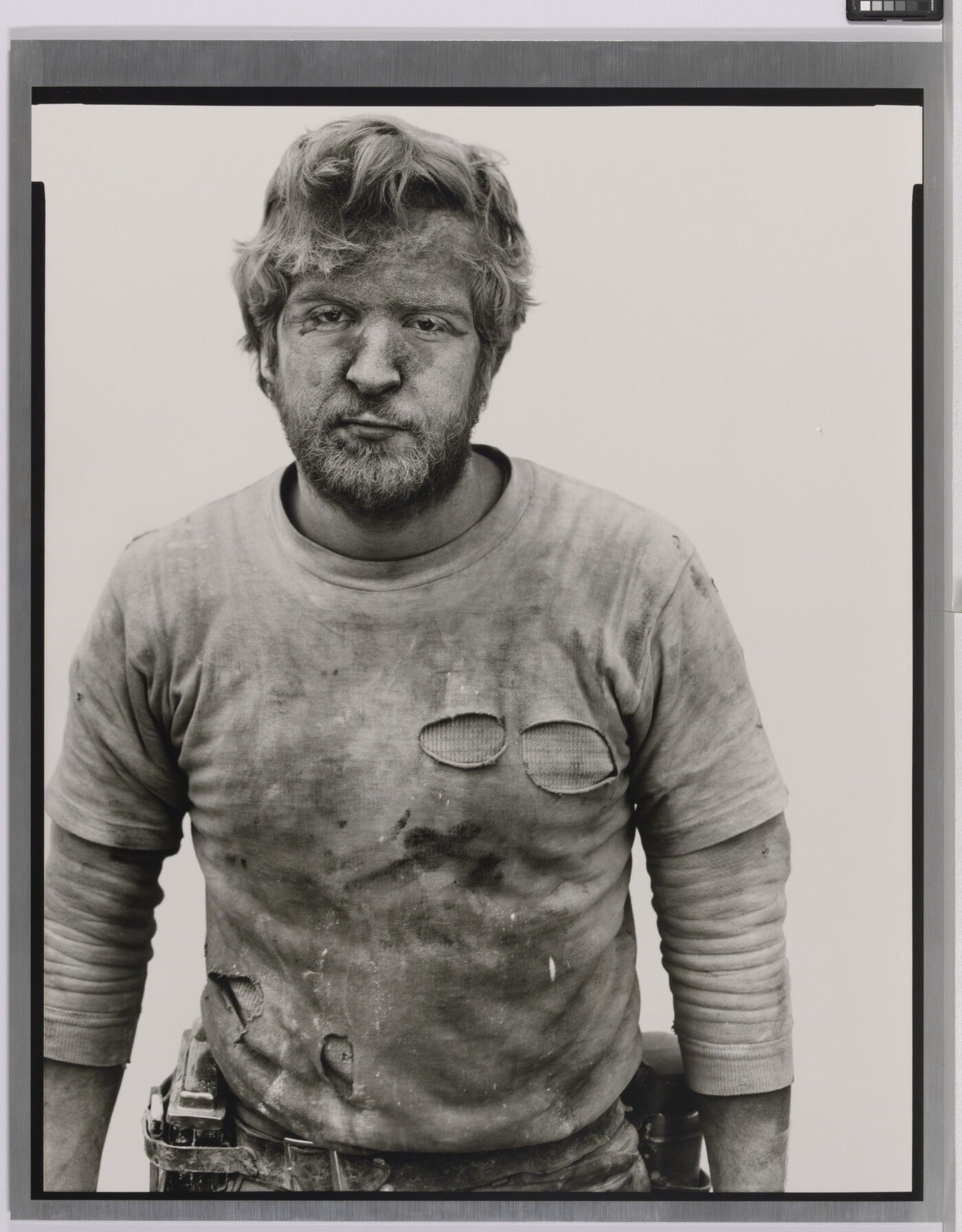 A black-and-white portrait photograph of a White man with messy hair and a beard wearing a dirty, ripped shirt.