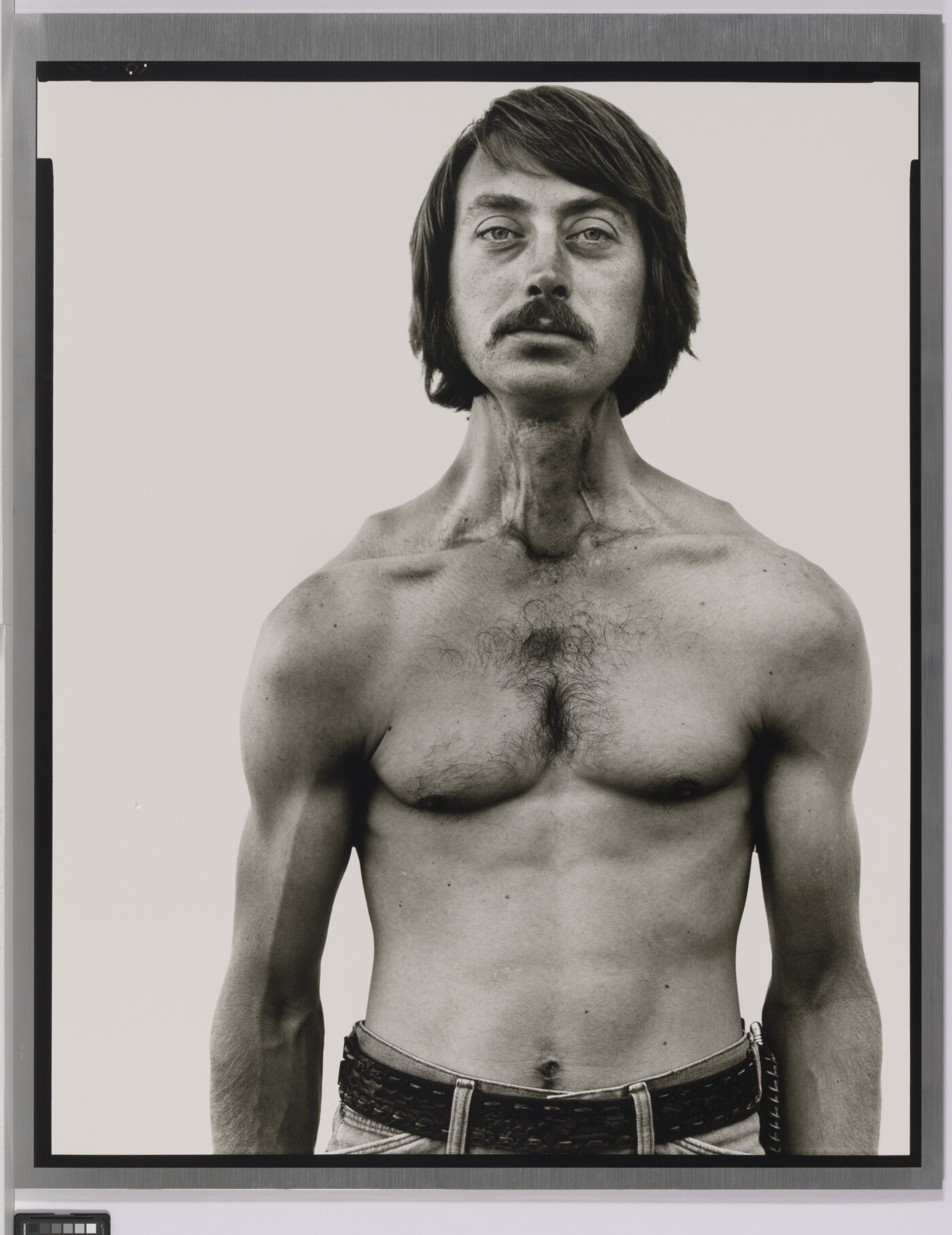 A black-and-white portrait photograph of a shirtless White man, with dark hair and a mustache, and thin but with muscled shoulders and arms.