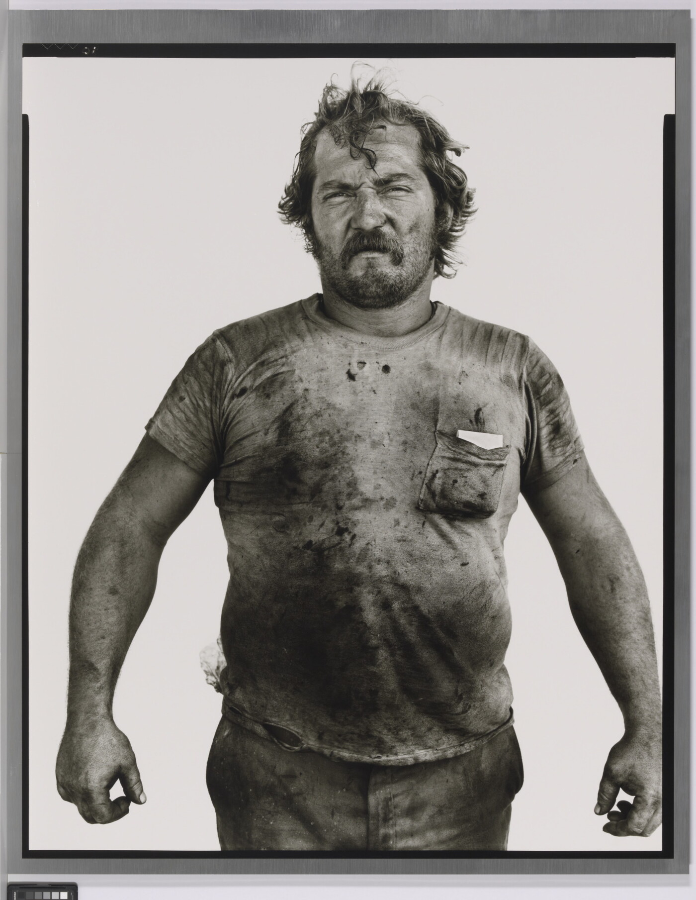 A black-and-white portrait photograph of a White man with messy hair and a beard wearing a dirty t-shirt and holding his arms out from his sides.