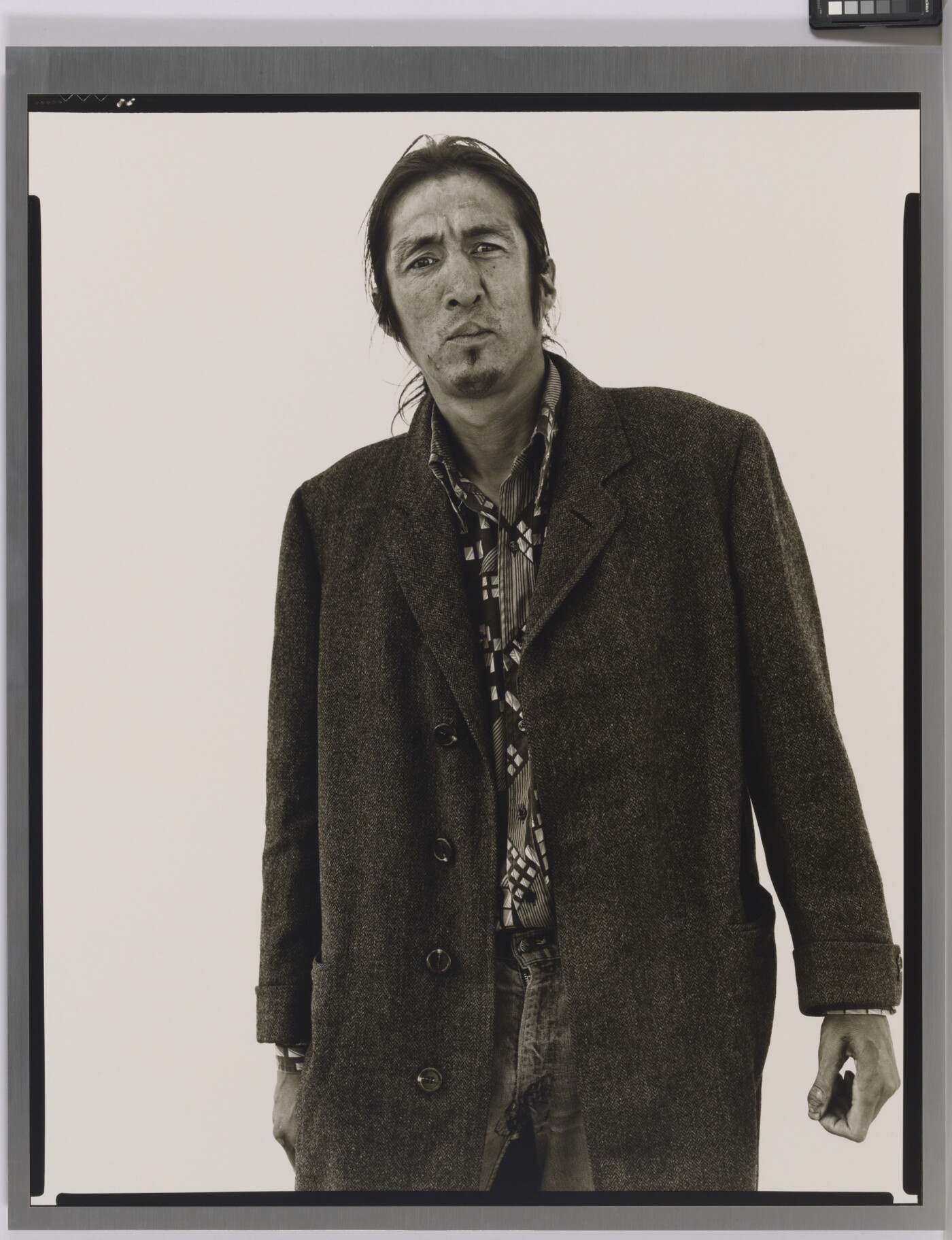 A black-and-white portrait photograph of a medium-skinned man with long hair, facial hear wearing a long coat.