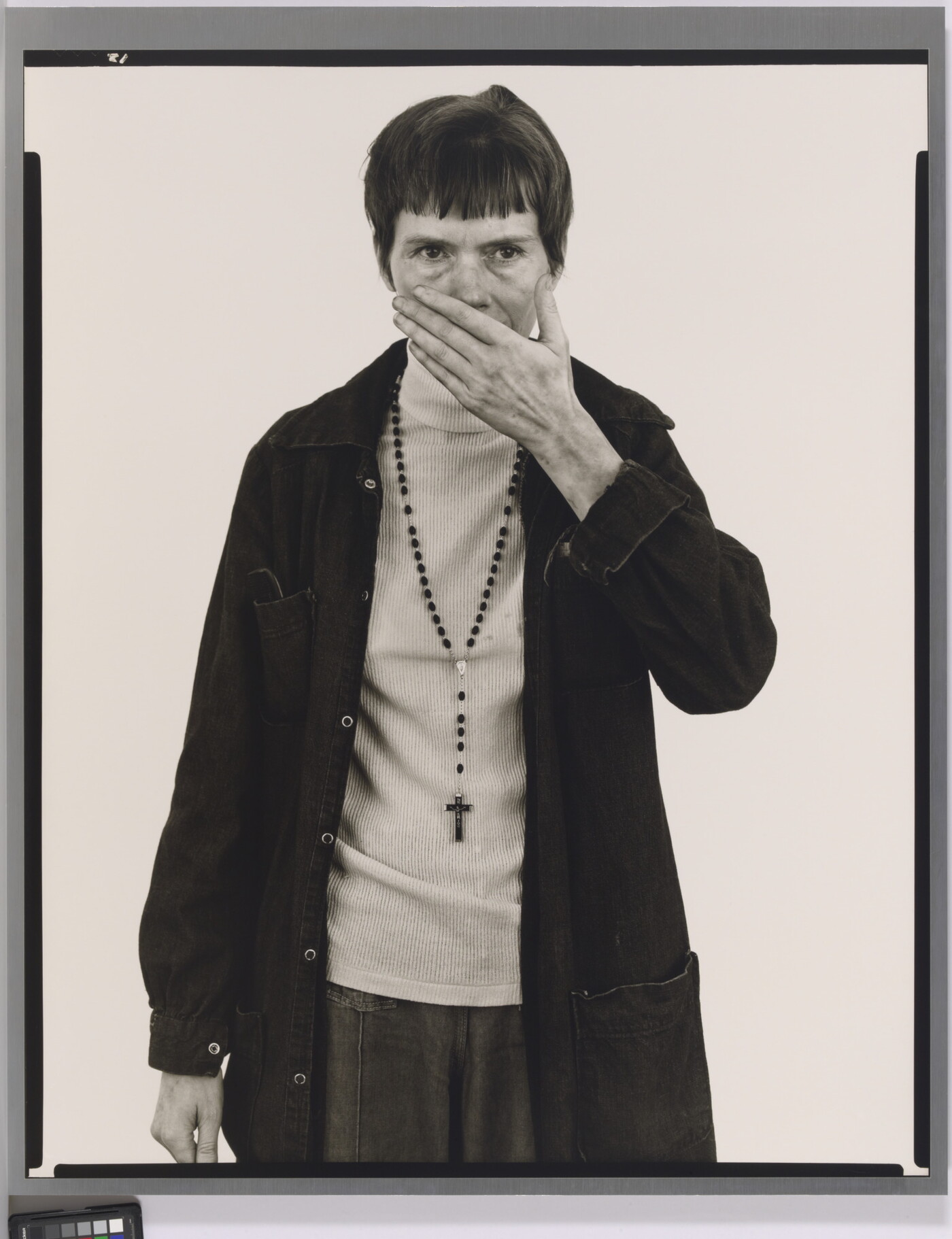 A black-and-white portrait photograph of a White woman with short, dark hair wearing a dark jacket and rosary beads around her neck holding one hand in front of her mouth and nose.