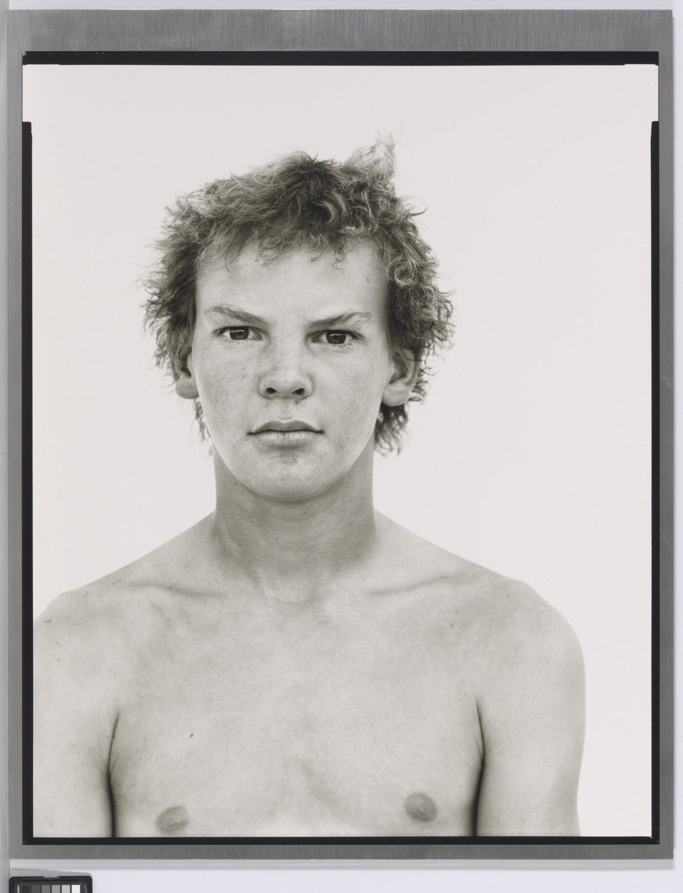 A black-and-white portrait photograph of a shirtless White teenage boy with messy hair.