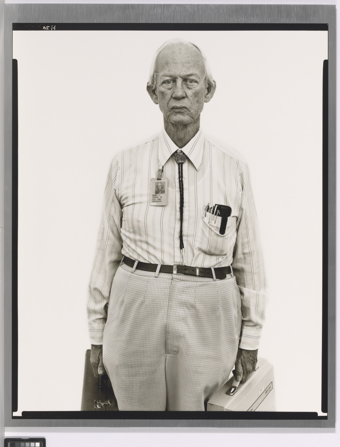A black-and-white portrait photograph of an older, mostly bald White man wearing a collared shirt, a bolo tie, and work badge attached to his collar, holding two briefcases.