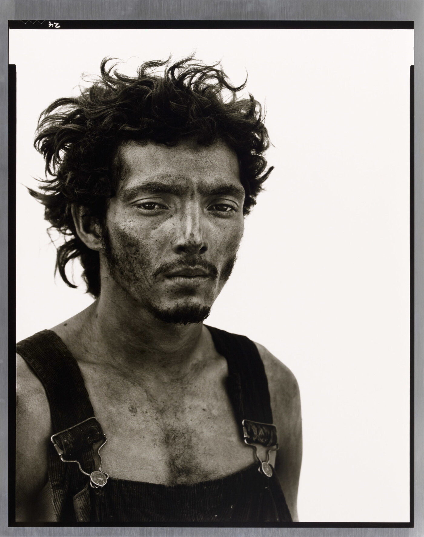 A black-and-white portrait photograph of a man from the chest up with messy hair, a dirty face, and overalls.