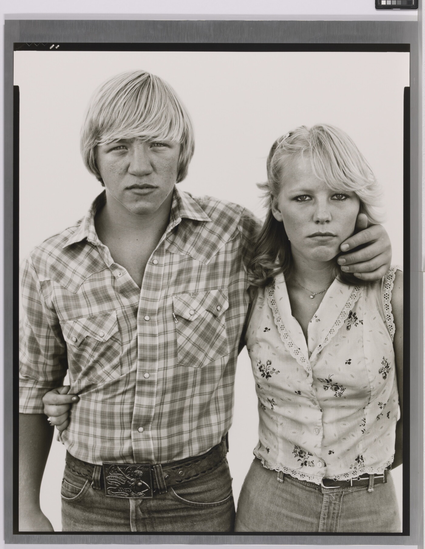 A black-and-white portrait photograph of a White teenage boy wearing a plaid button-down shirt, his arm around the shoulders of a White teenage girl wearing a sleeveless shirt.