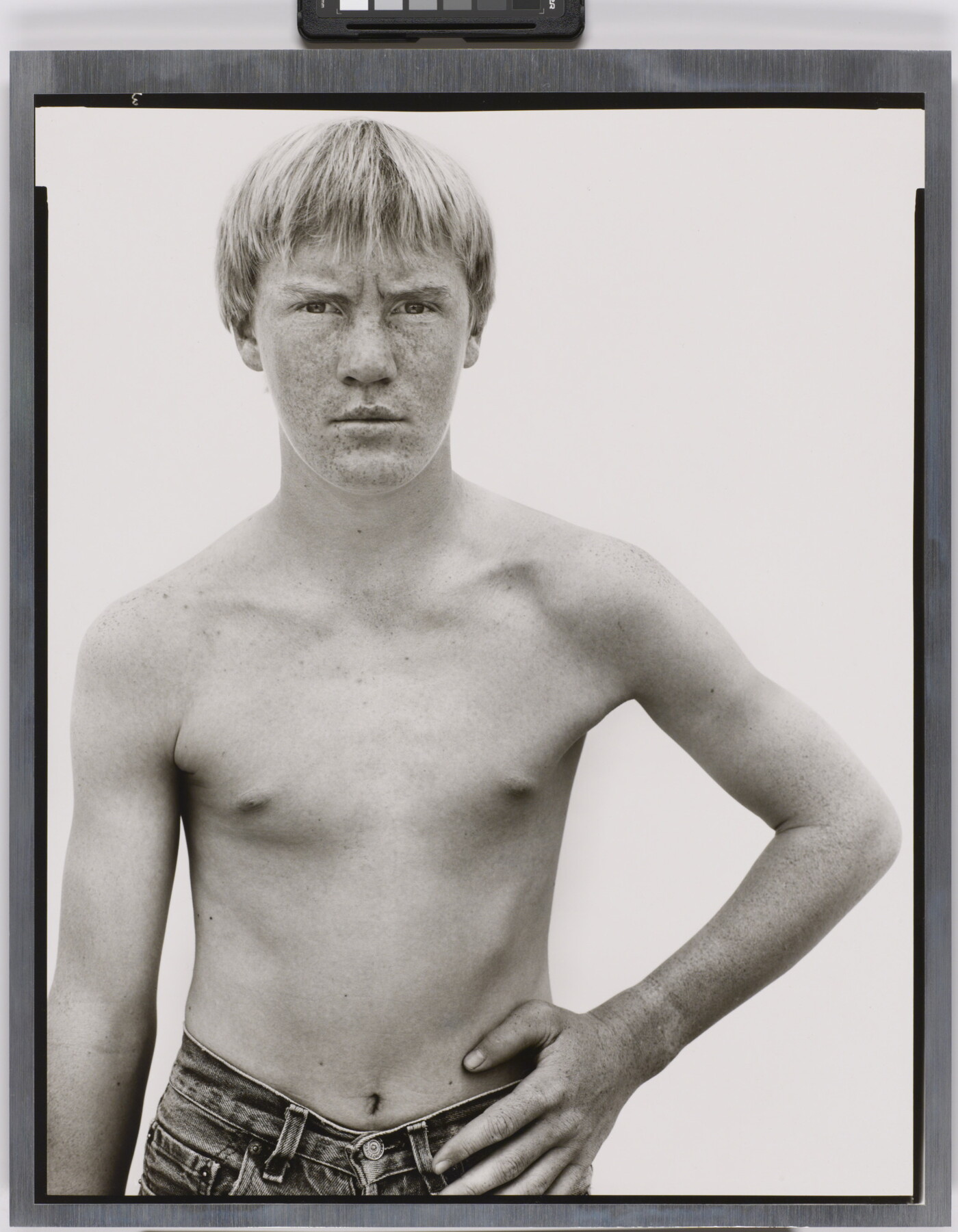 A black-and-white photograph of a shirtless White boy with light hair and a freckled face standing with one hand on his hip.