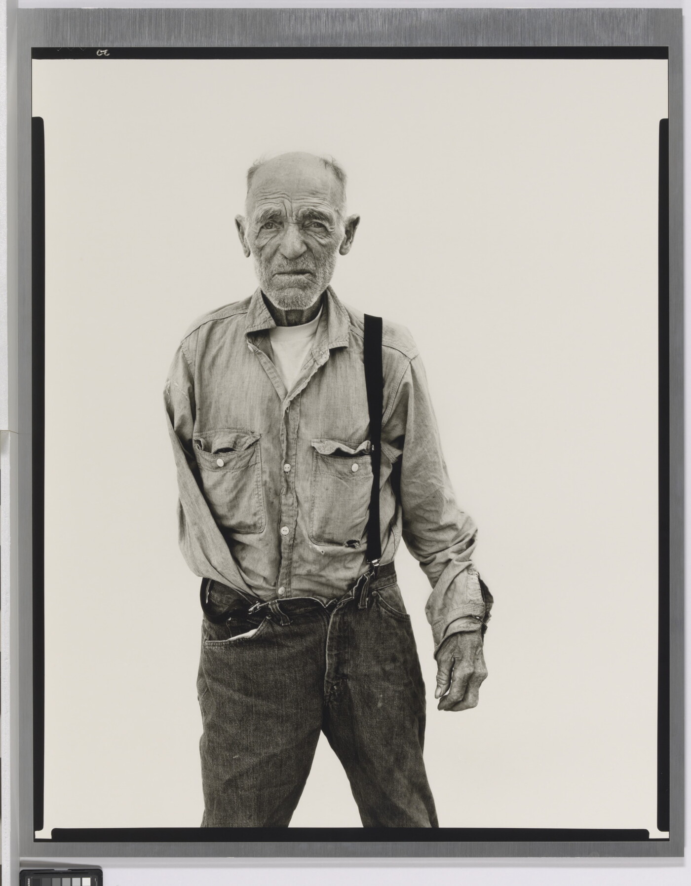 A black-and-white portrait photograph of an older, balding White man with one arm wearing a button-down shirt and jeans held up with one suspender.