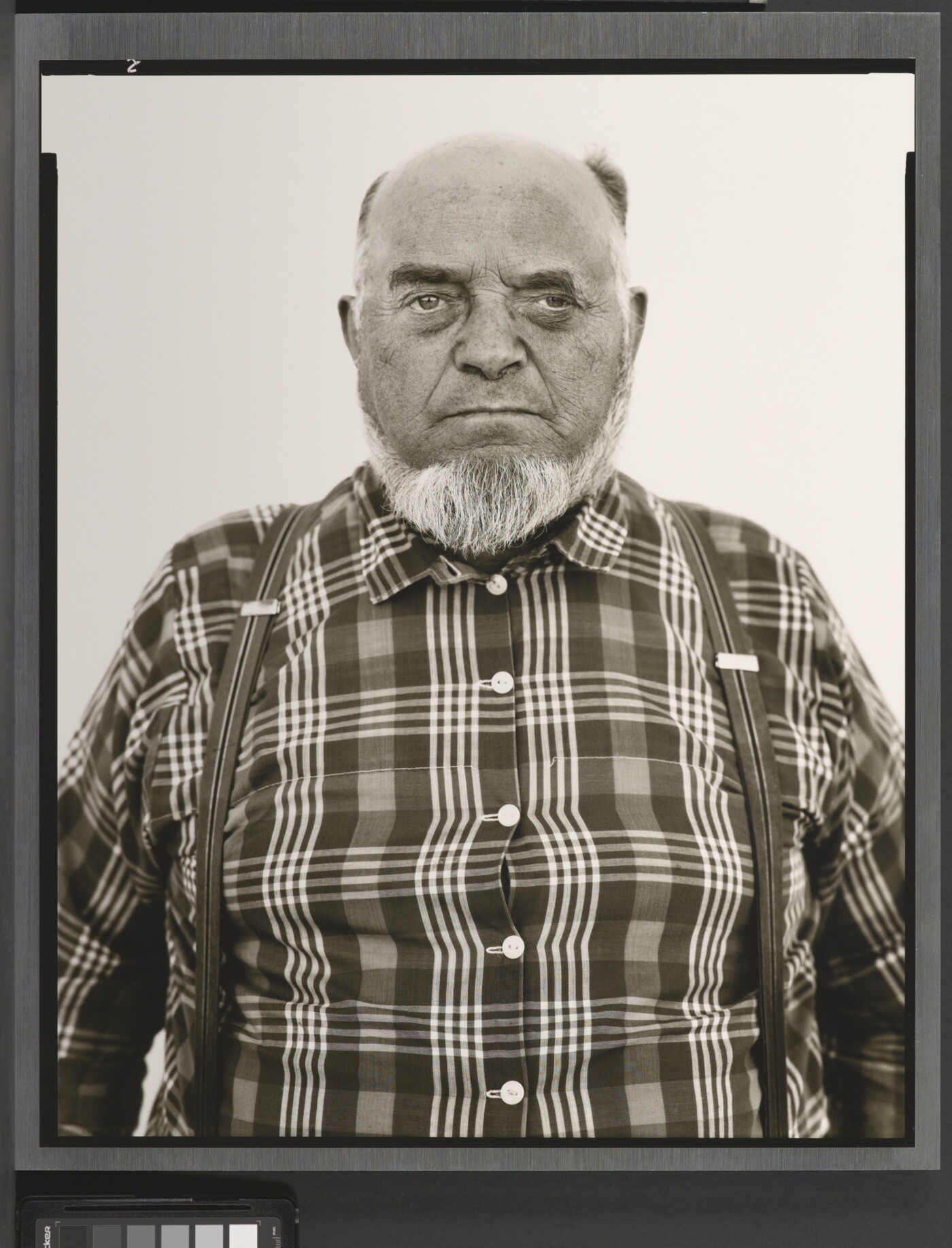 A black-and-white portrait photograph of an older, balding White man with a gray beard wearing a plaid button-down shirt and suspenders.