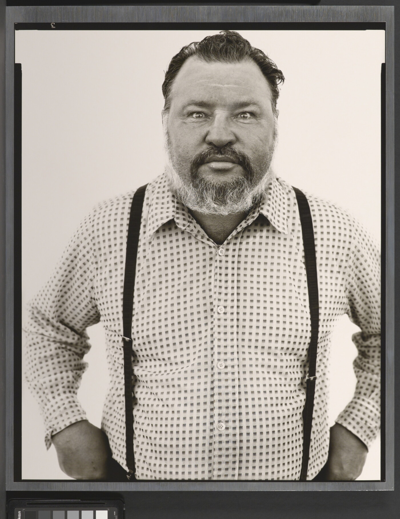 A black-and-white portrait photograph of a middle-aged White man with a receding hairline and salt-and-pepper beard wearing a checked button-down shirt and suspenders.