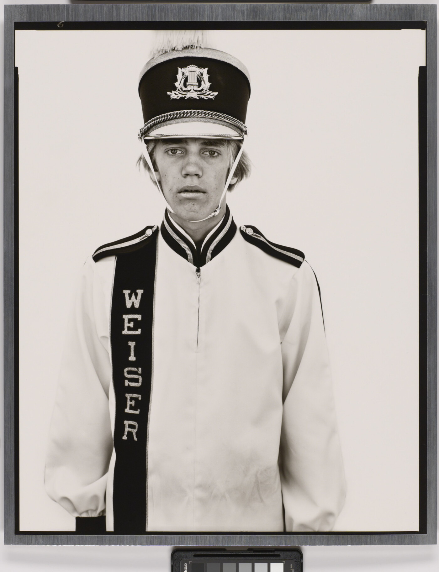 A black-and-white portrait photograph of a White teenage boy wearing an oversized, white, marching-band hat and uniform.