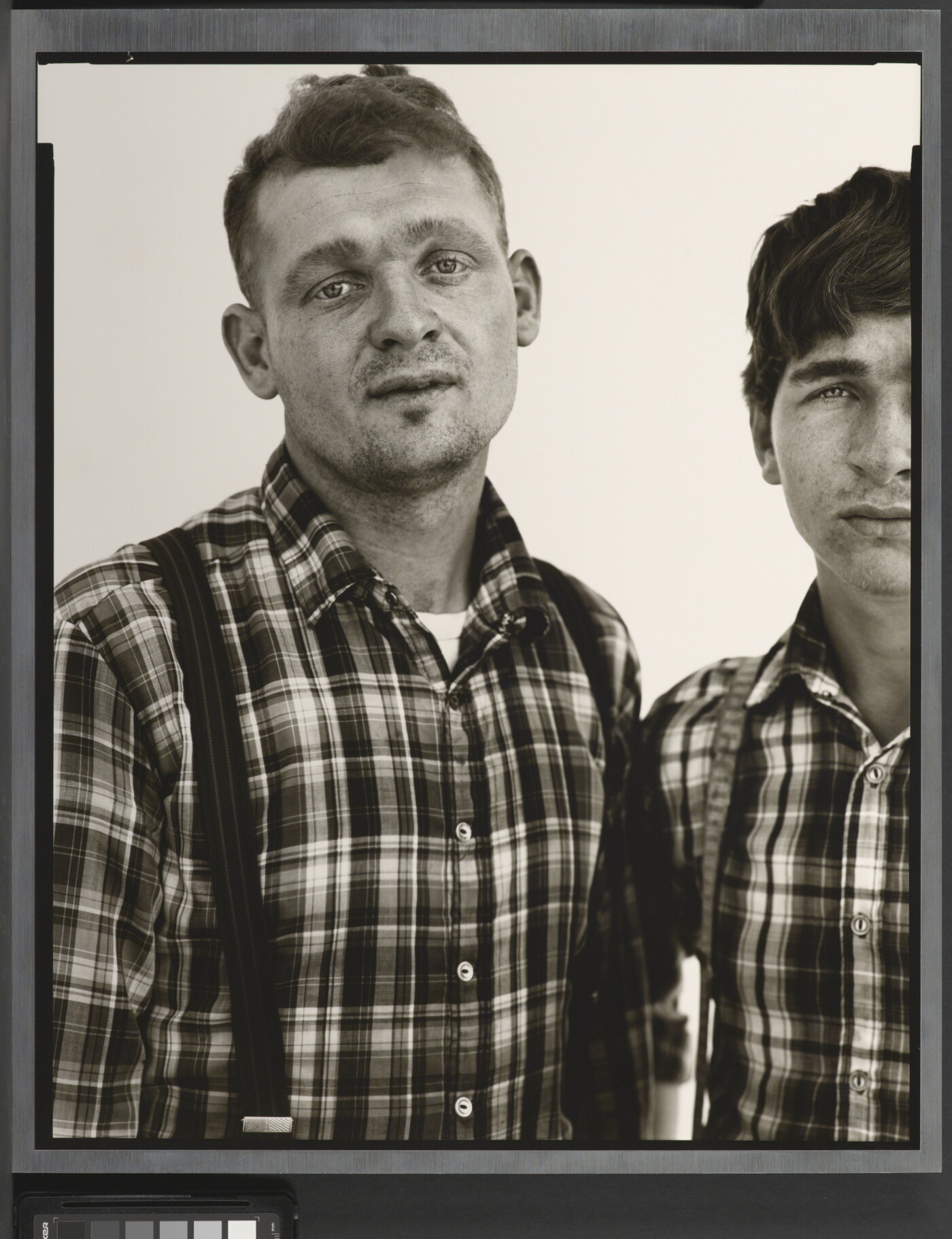 A black-and-white portrait photograph of two White men, both in similar plaid shirts and suspenders, standing shoulder to shoulder.
