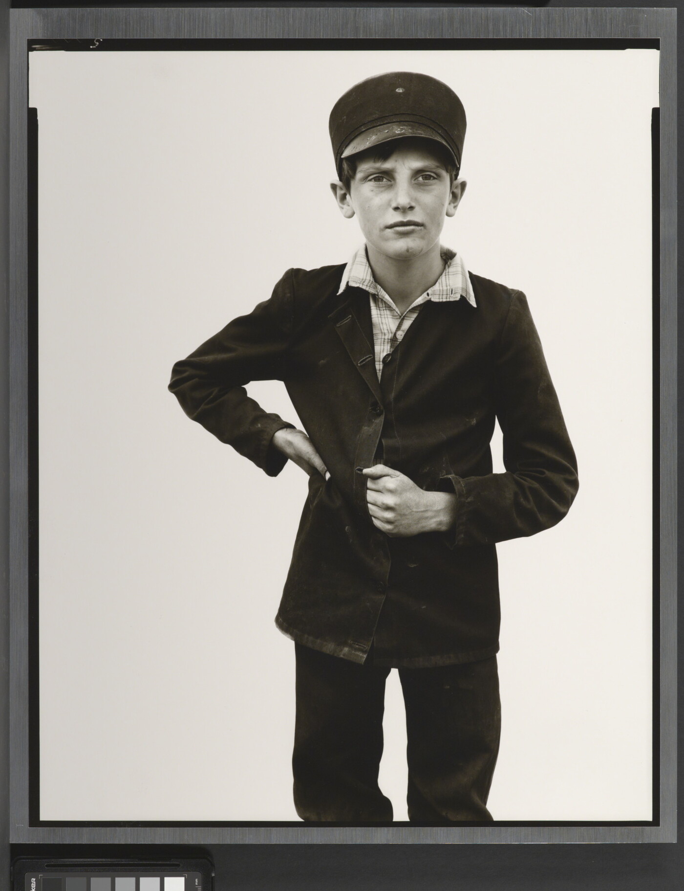 A black-and-white portrait photograph of a White boy wearing a Breton cap, a dark button-up jacket with one hand on his waist and the other tucked in between the buttons.