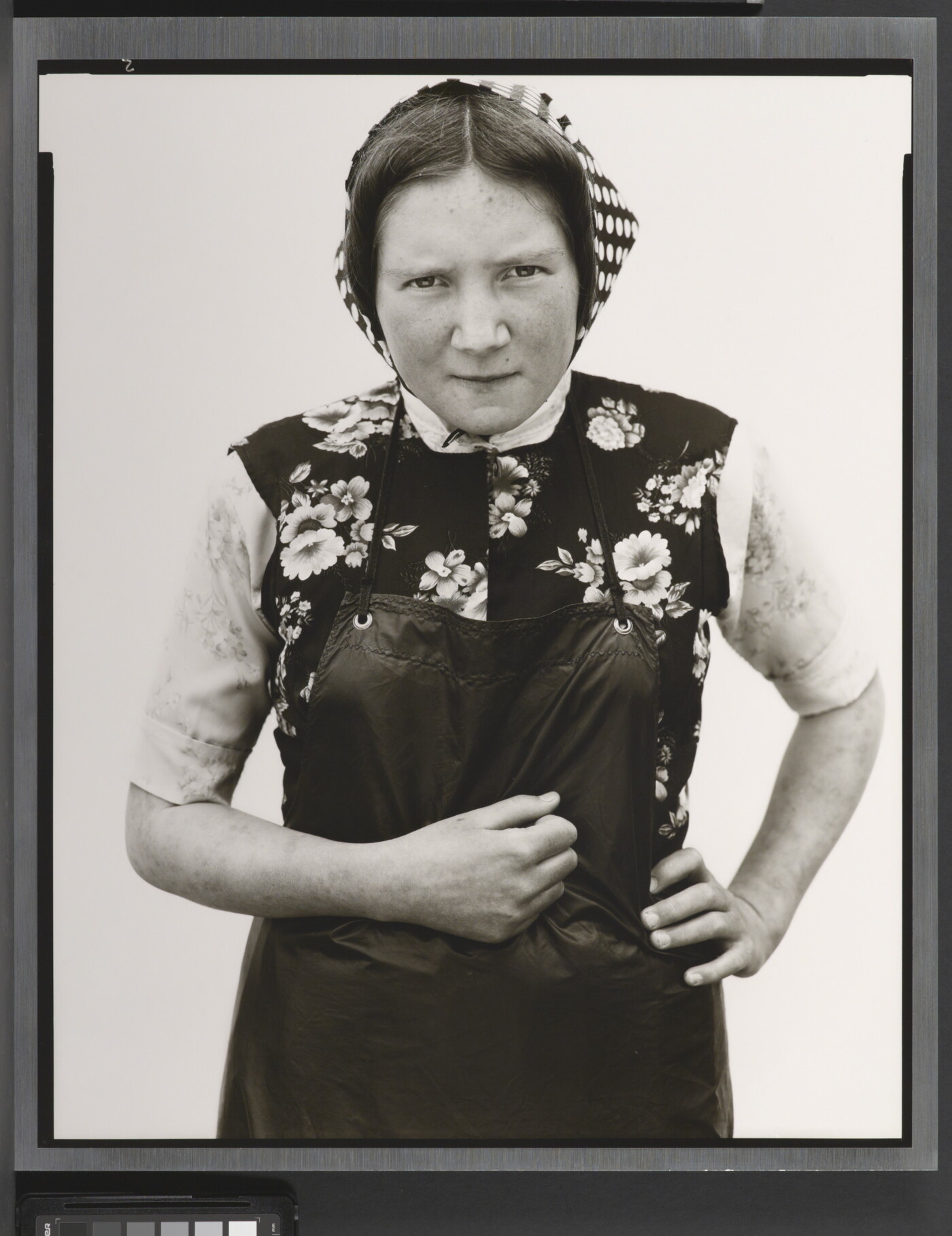 A black-and-white portrait photograph of young White woman wearing a checked kerchief, flowered vest, and dark apron.