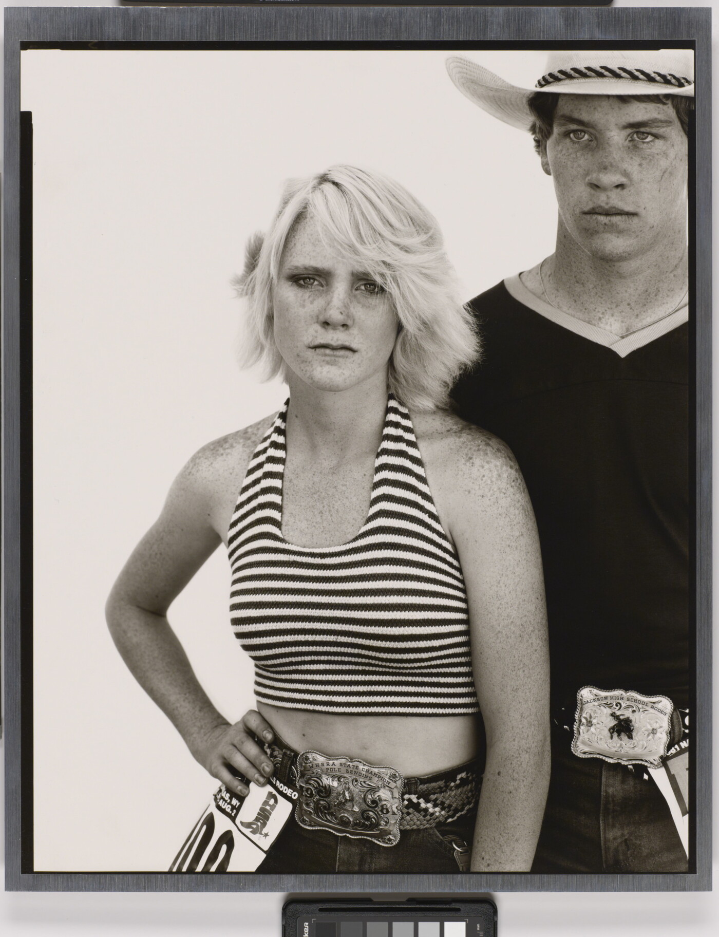 A black-and-white portrait photograph of a young White woman wearing a striped halter top, one hand on her hip, and a young White man wearing a cowboy hat and V-neck t-shirt, both wearing large buckles on their belts.