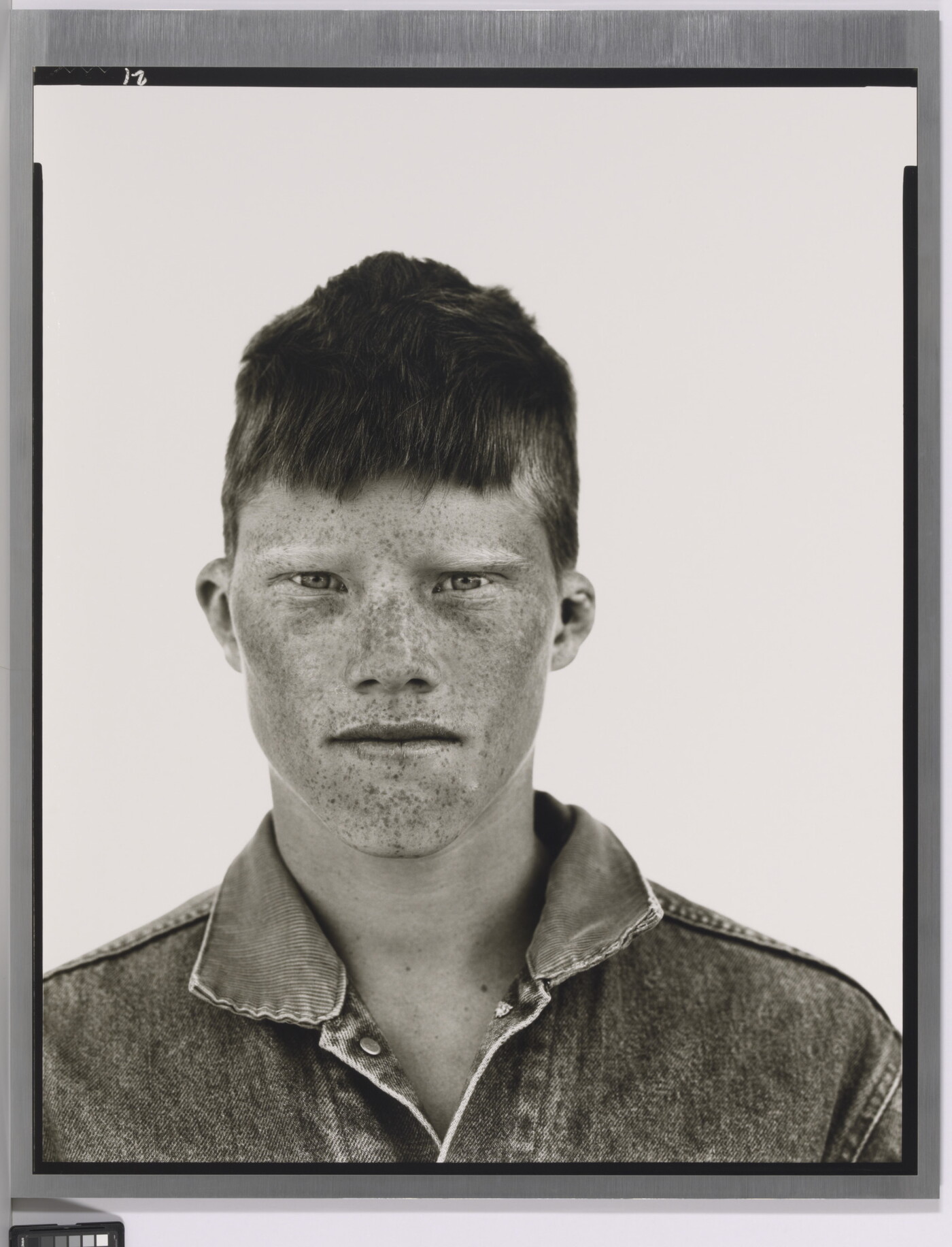 A black-and-white portrait photograph of a White teenage boy, face covered in freckles, short cropped hair, wearing a denim jacket.