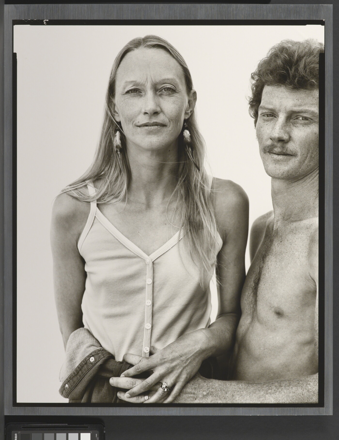 A black-and-white portrait photograph of a thin White woman with long hair wearing camisole as a shirtless White man with curly hair and a moustache holds her waist.