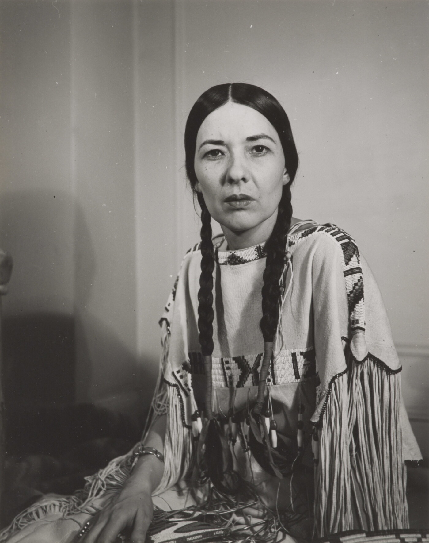A black-and-white photograph of a seated Indigenous woman, hair in braids, gazing into the camera. 