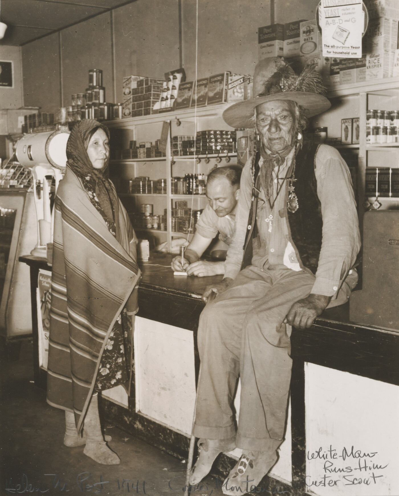 A sepia-toned photograph of a Indigenous man sitting on a counter, an Indigenous woman wrapped in a blanket standing at the counter, and a White man behind the counter.