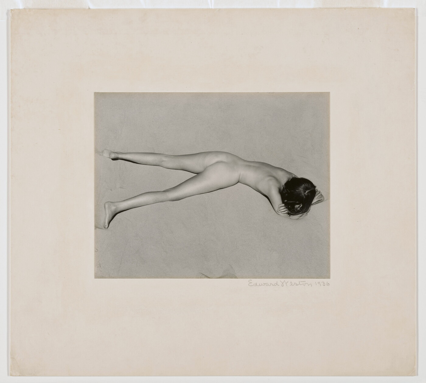 A black-and-white photograph of a nude, fair-skinned woman lying facedown in the sand with her head resting on her crossed arms.