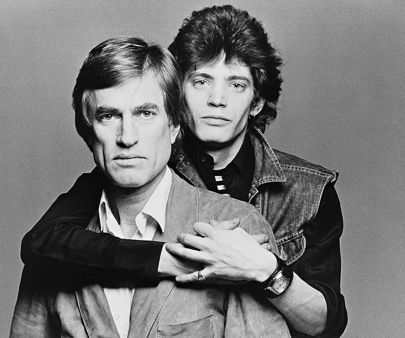 A black-and-white studio portrait photograph of two White men, one standing behind the other with his arms wrapped around the other&#039;s shoulders.