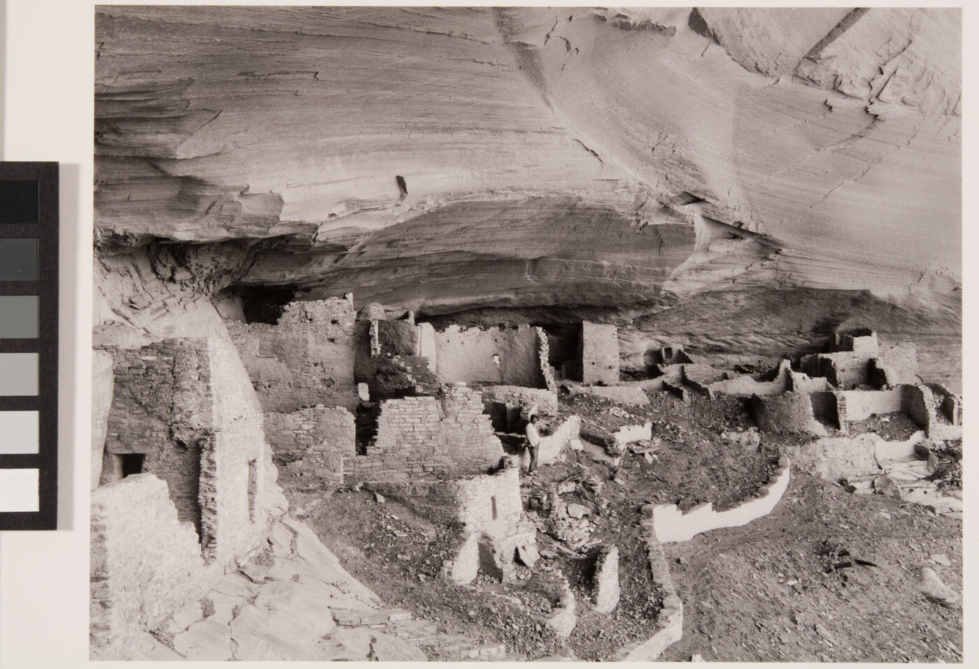 A black-and-white photograph of adobe ruins under an overhanging cliff ledge.