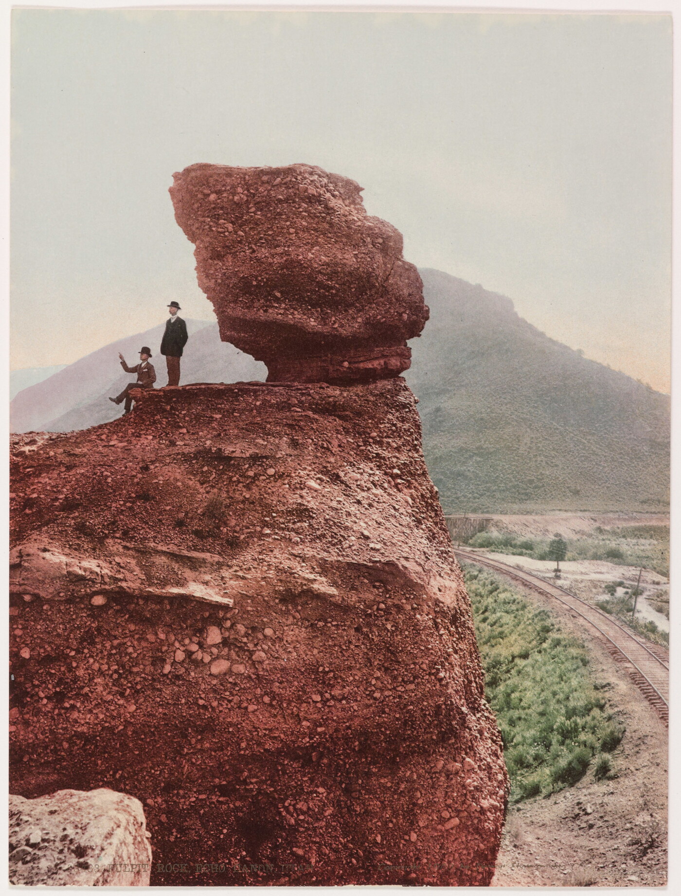 A color photograph of two men, one seated and one standing, next to a large rock formation with a mountain in the background.