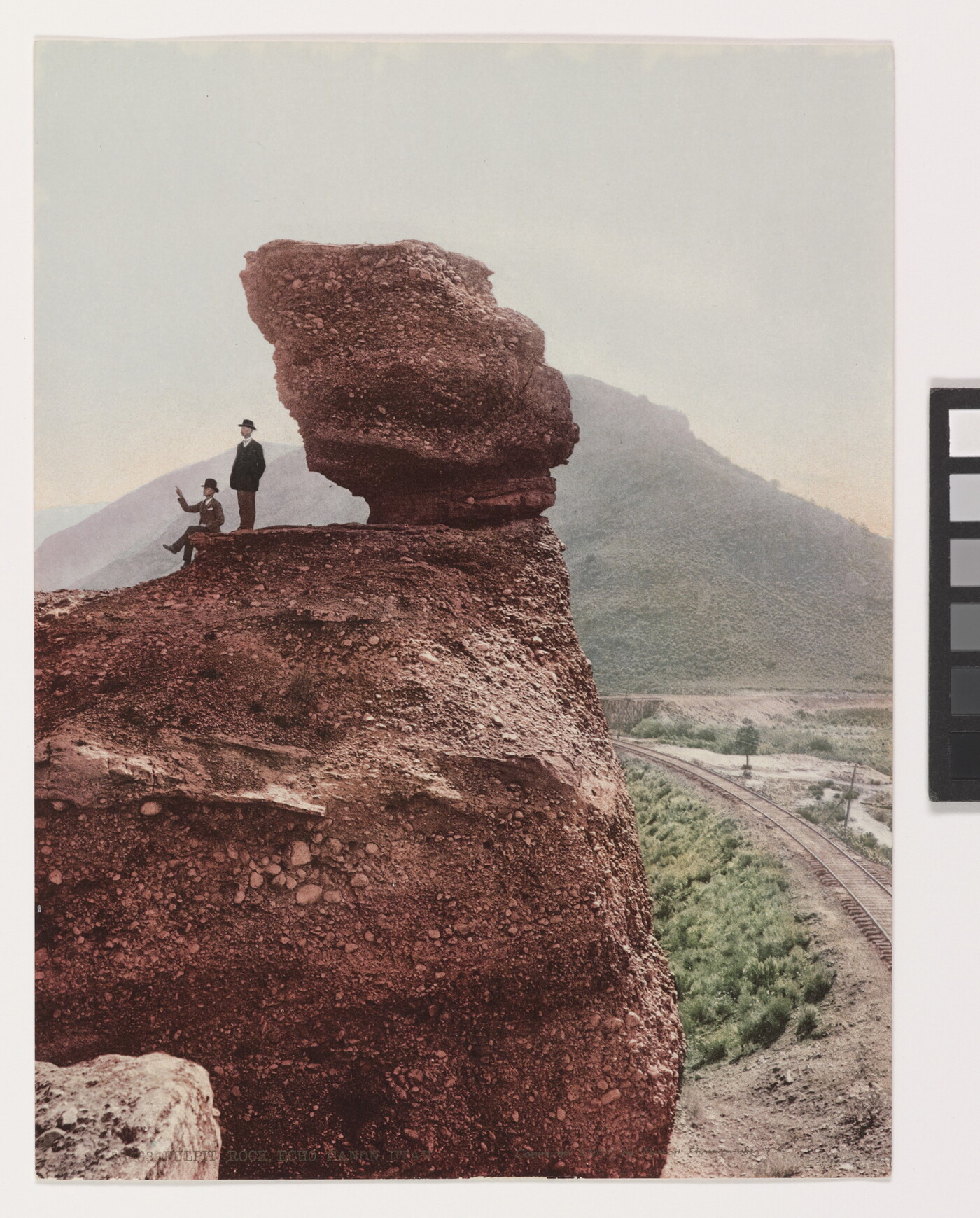 A color photograph of two men, one seated and one standing, next to a large rock formation with a mountain in the background.