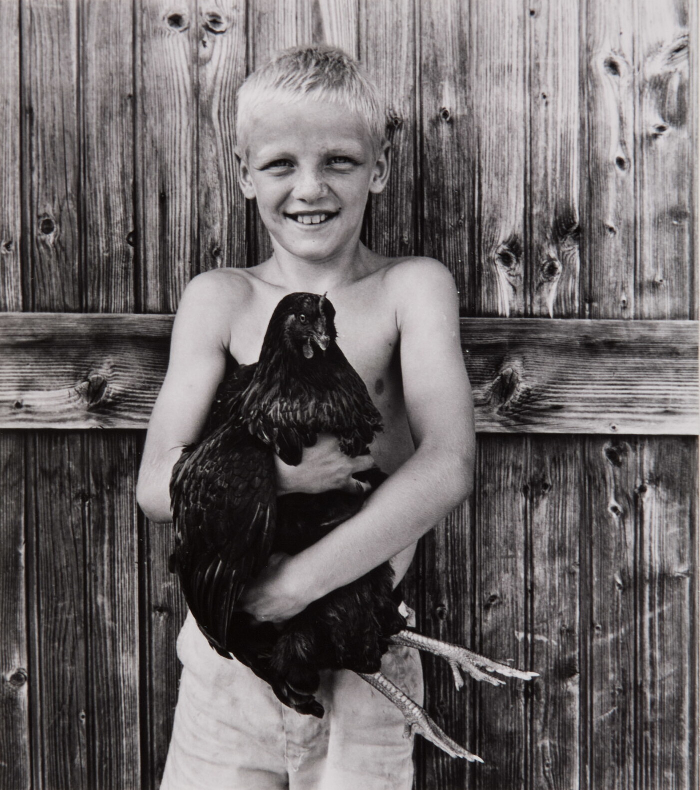 A black-and-white photograph of a smiling, shirtless White boy holding a chicken to his chest.