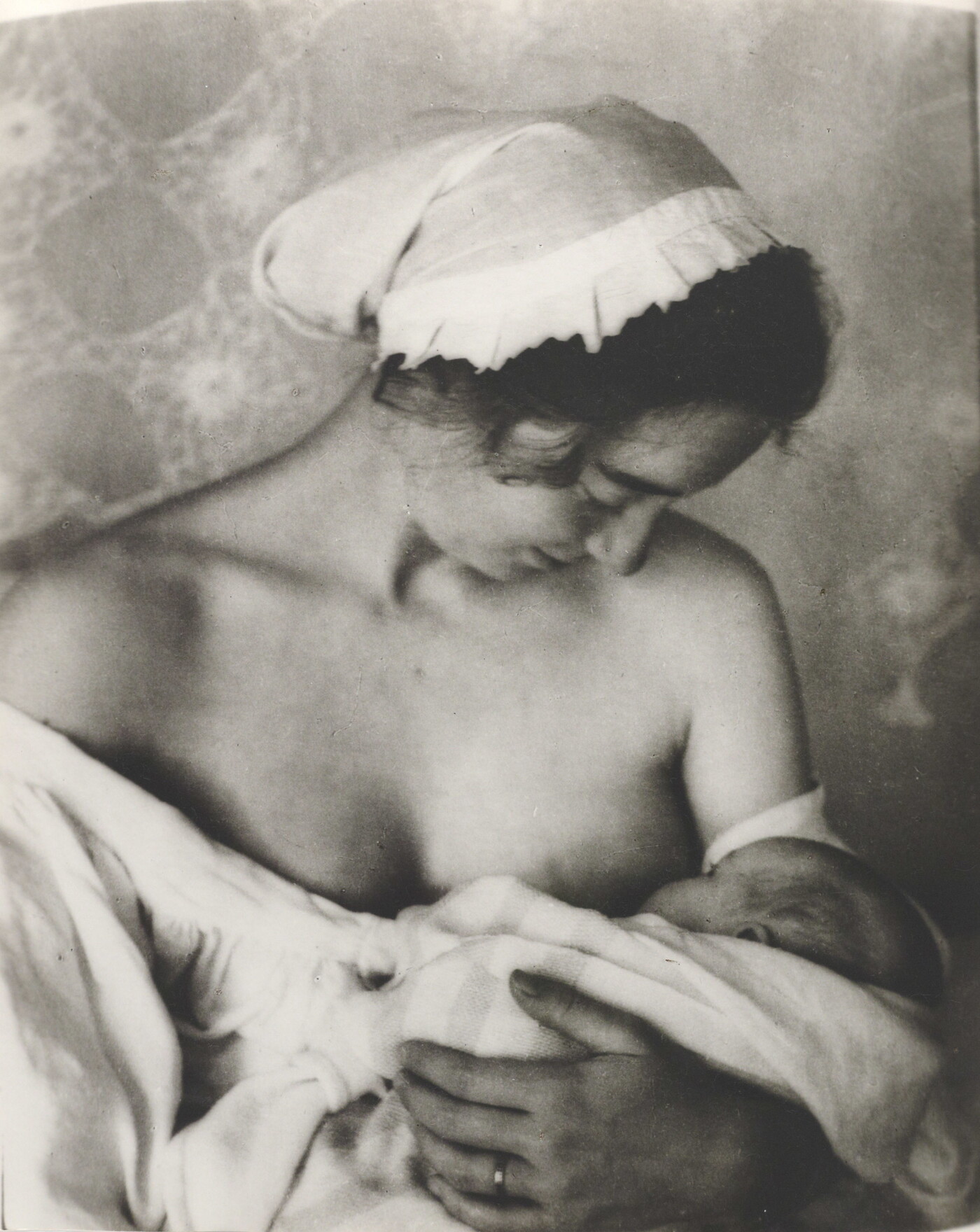 A black-and-white photograph of a White woman in a nightcap and white flowing blouse nursing an infant.