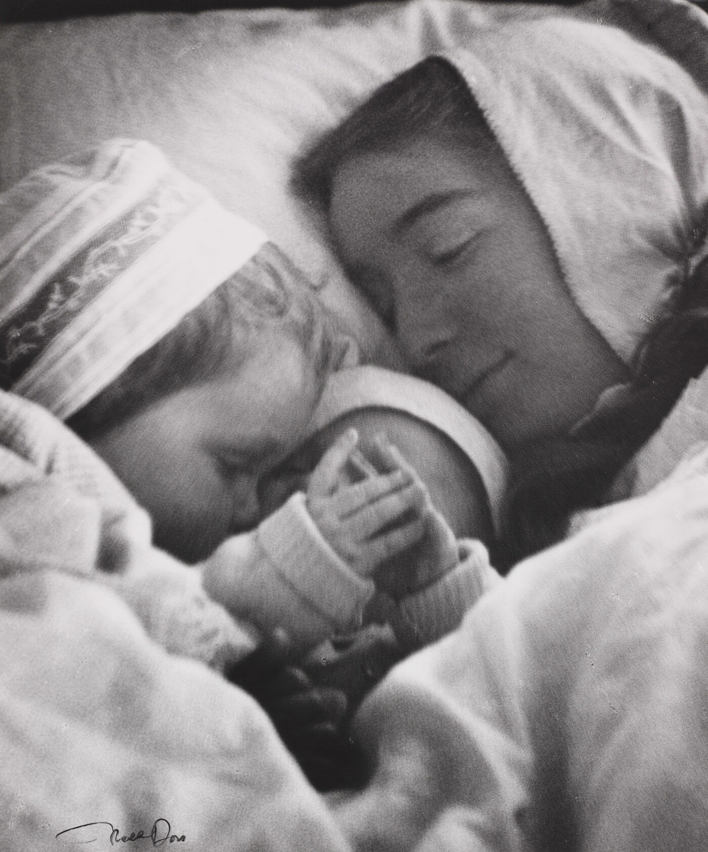 A black-and-white photograph of a White woman in a nightcap cuddling in bed with a toddler and an infant.