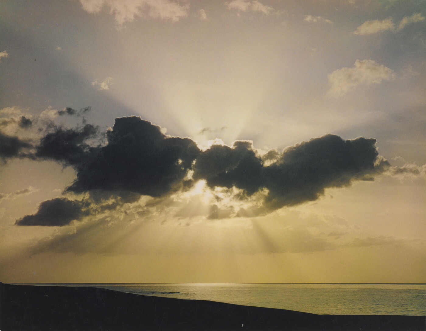 A color photograph of a sun peeking from behind clouds over a calm beach.