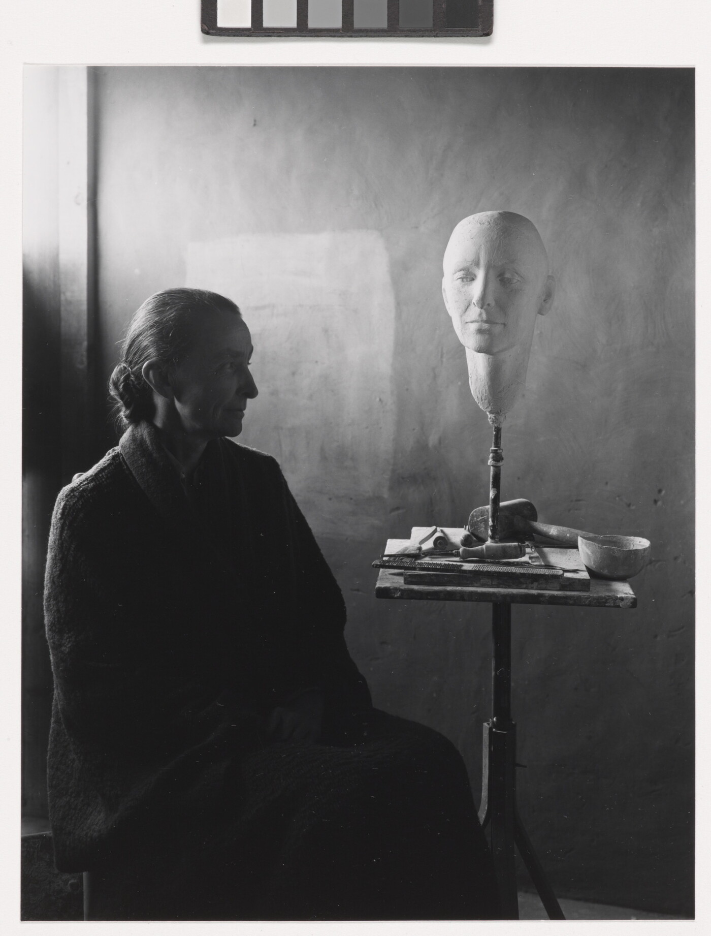 A black-and-white portrait photograph of a seated older White woman wearing a dark, floor length dress; a white face mask sits on a stand on a cluttered end table.