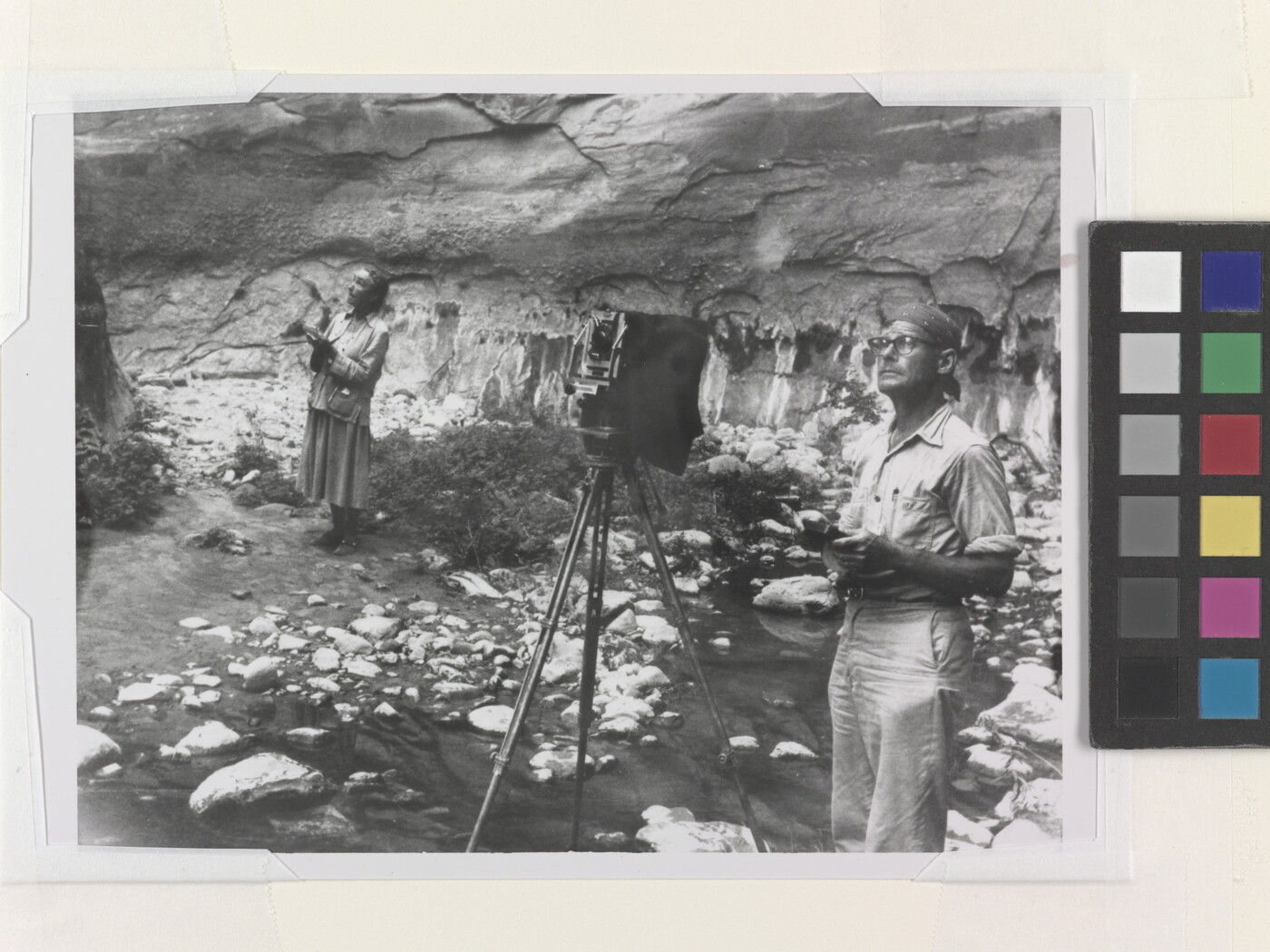 A black-and-white photograph of a White woman with a sketchbook and a White man with a camera next to a rock wall, both looking at something out of the frame.