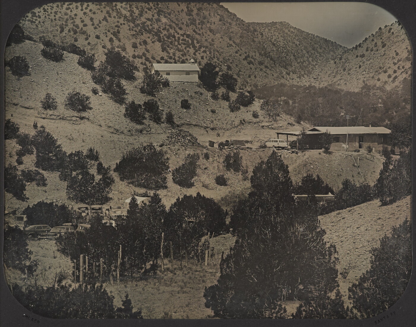 A black-and-white photograph of several buildings on a mountainside dotted with pine trees.