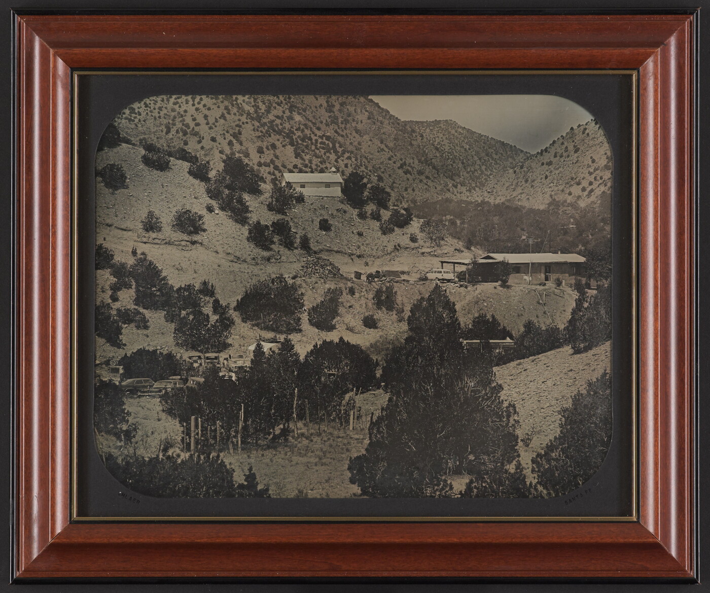 A black-and-white photograph of several buildings on a mountainside dotted with pine trees.