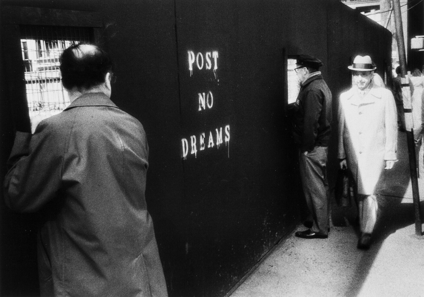 A black-and-white photograph of men looking through the windows of a construction wall; the wall has &quot;Post no dreams&quot; painted on it.