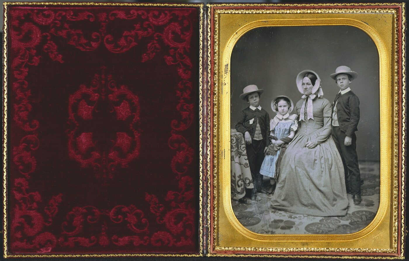 A hand-painted black-and-white formal portrait photograph in a gold frame of a White woman sitting with two boys and a girl standing around her.