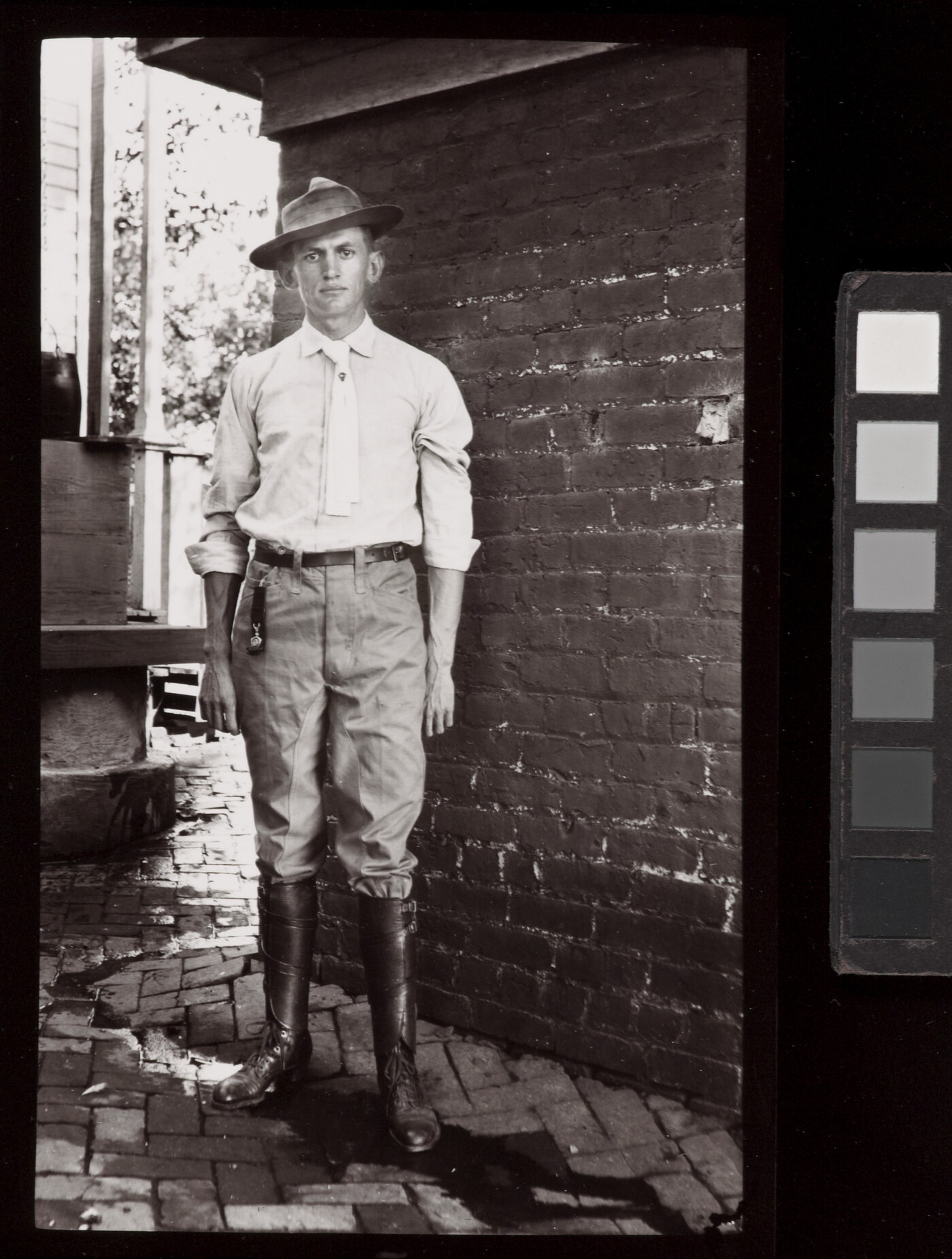 A black-and-white photograph of a young White man in a brimmed hat, light shirt and tie, pants, and tall boots standing in front of brick wall.