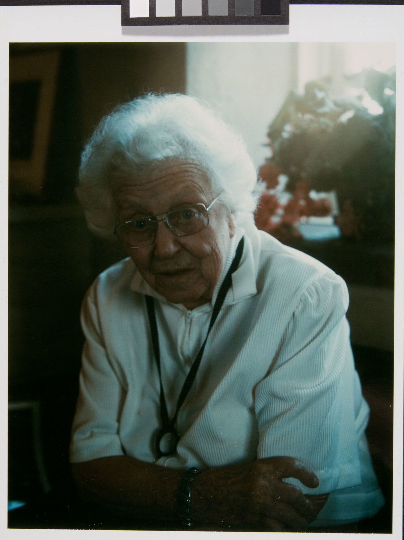 A color photograph of an older White woman with white hair, glasses, and a white blouse leaning forward with her arms crossed.