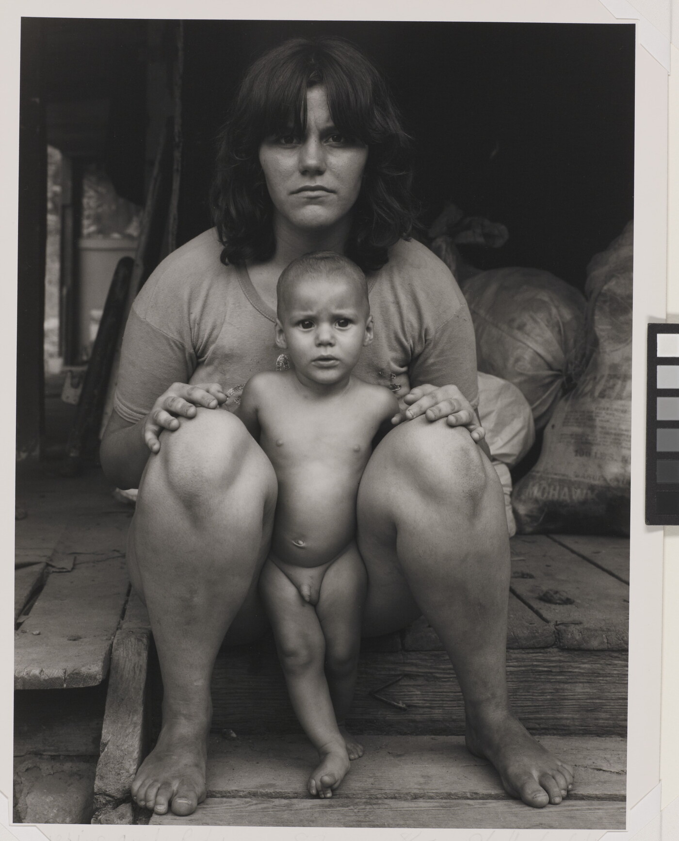 A black-and-white photograph of an expressionless White woman sitting on a wood step with a naked baby standing between her legs.