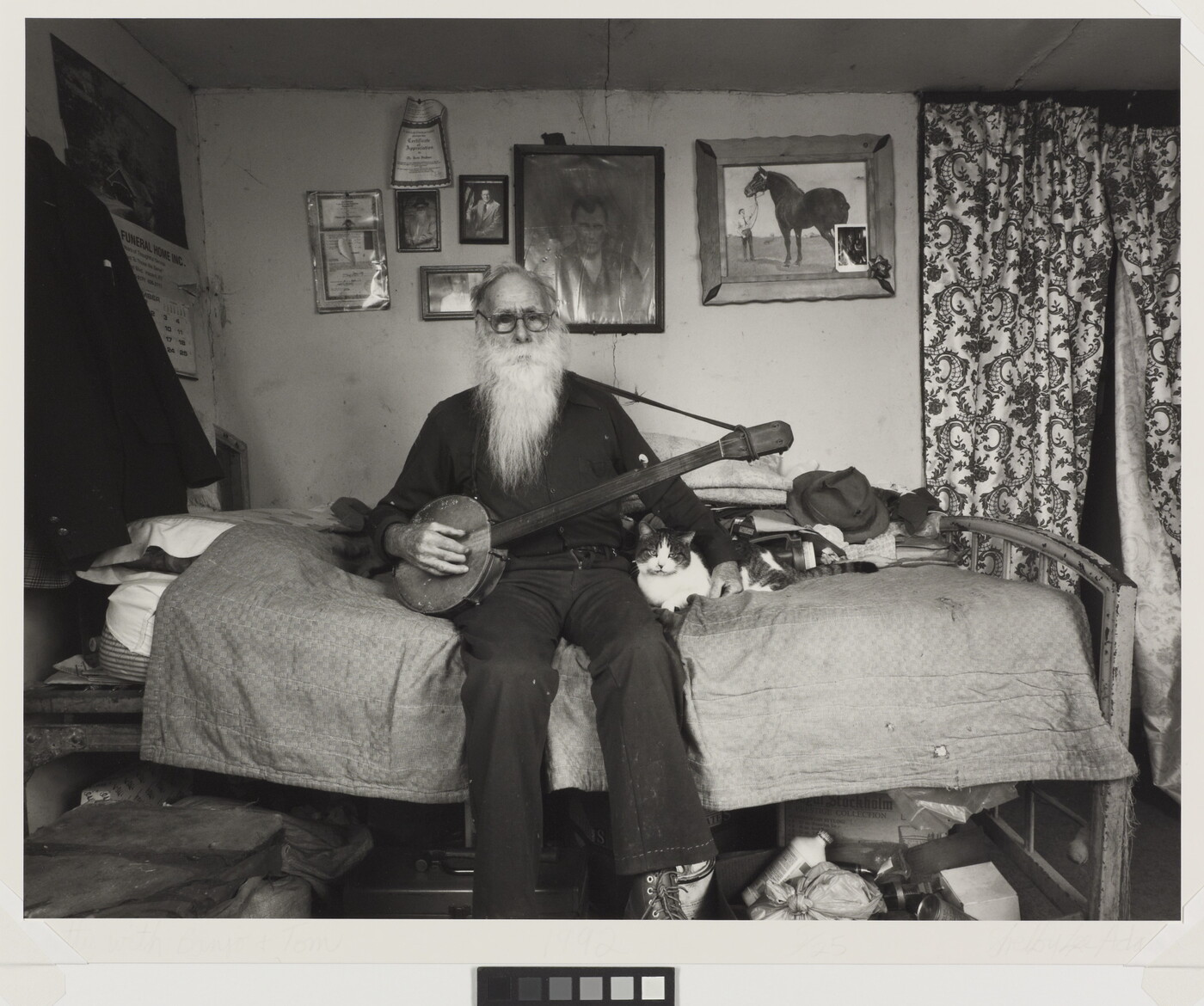 A black-and-white photograph of an elderly White man with a long white beard sitting on a bed in a cozy bedroom holding a banjo with one hand, the other hand around a cat lying next to him.