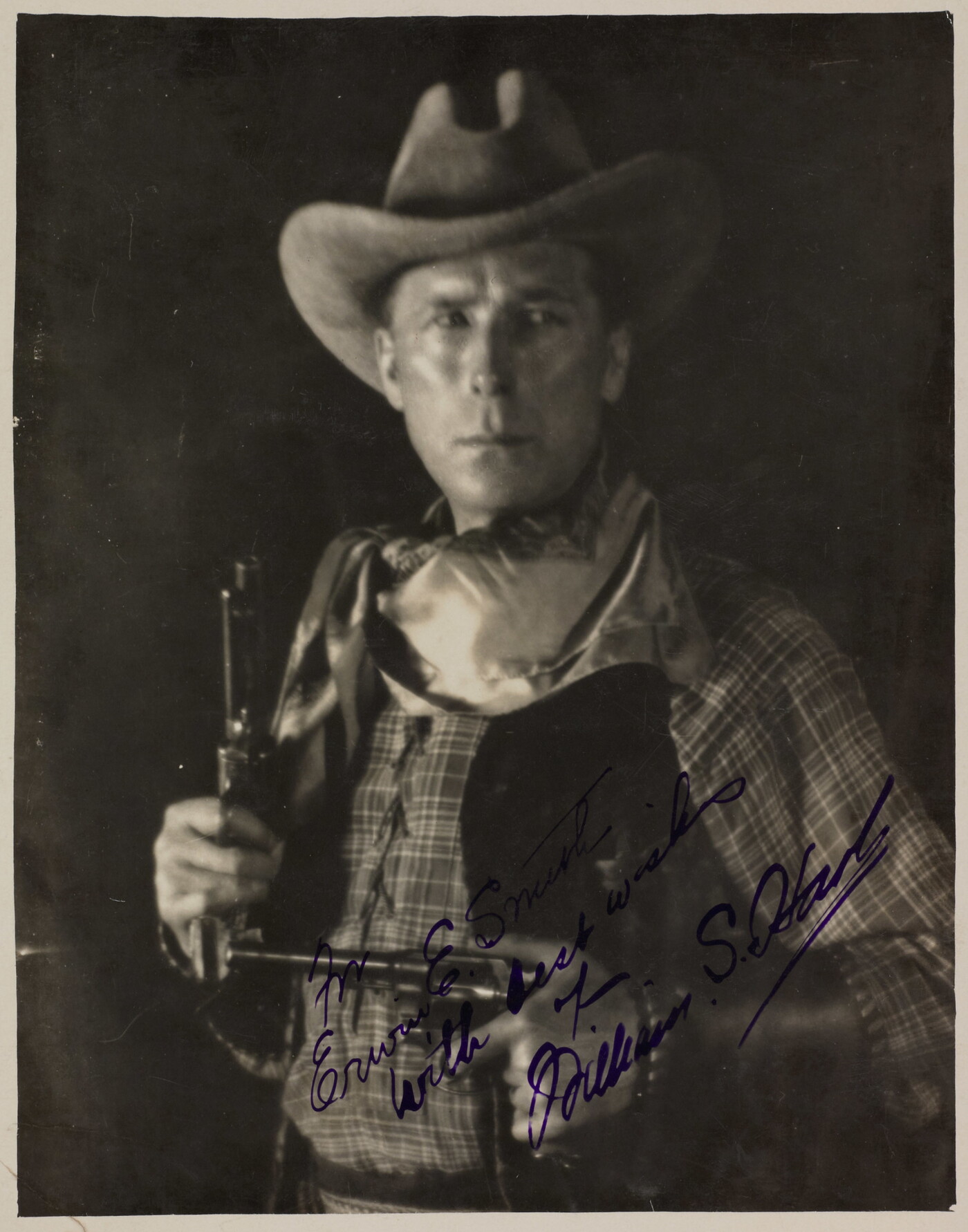 A black-and-white photograph of a White man in western attire holding a pistol in each hand, an autograph in blue in across the bottom.
