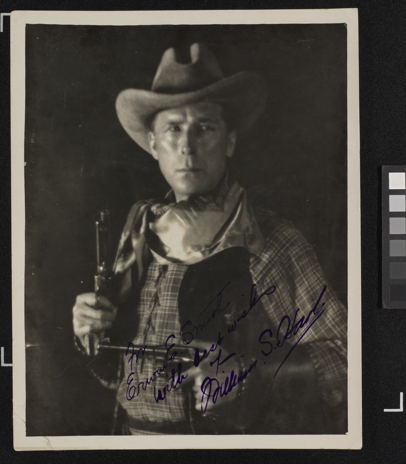 A black-and-white photograph of a White man in western attire holding a pistol in each hand, an autograph in blue in across the bottom.
